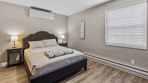 Ein Schlafzimmer mit Bett, Nachttischen, einer Wand-Klimaanlage, einem Fenster mit Jalousien und einem motivierenden Zitat an der Wand.