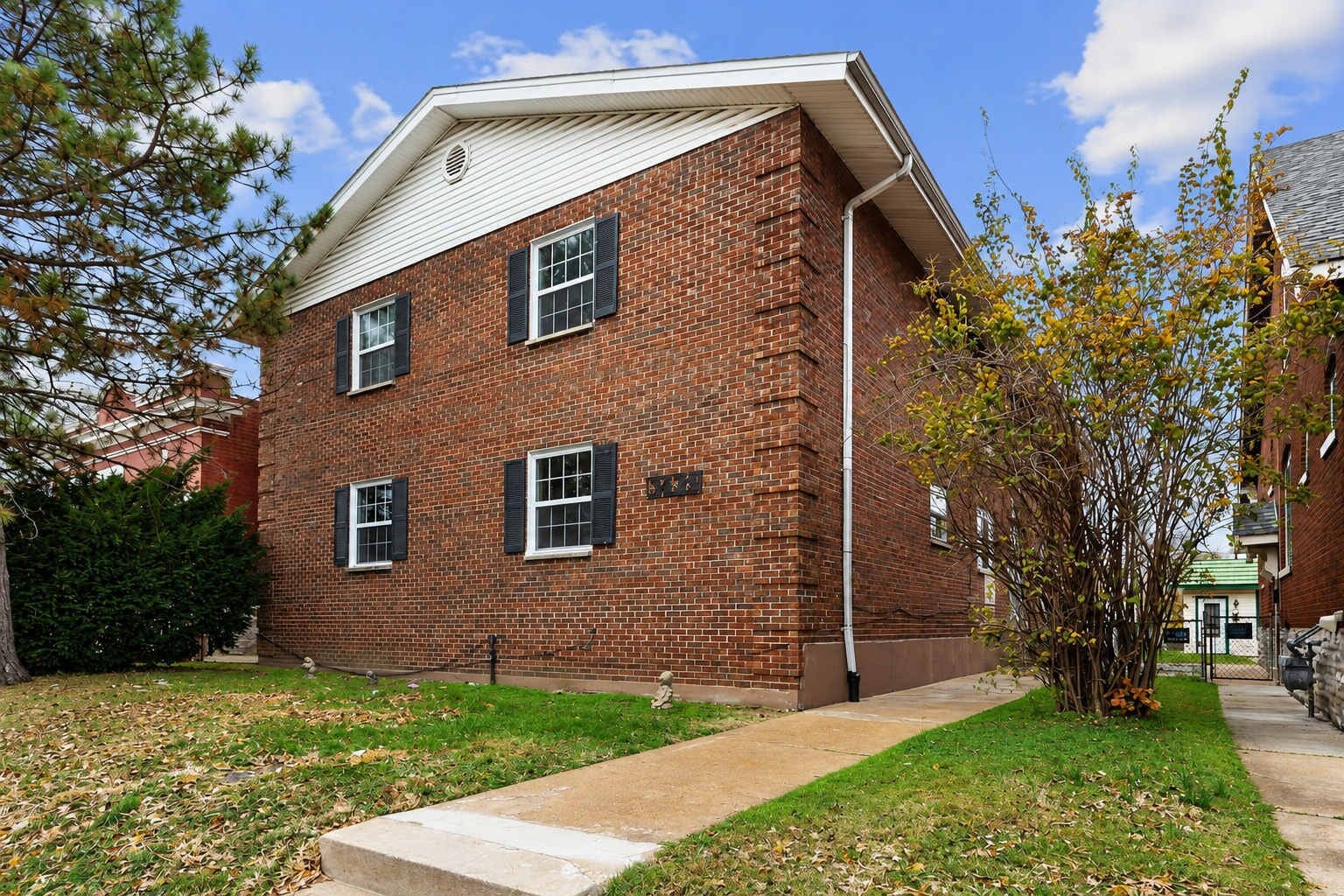 A two-story brick building with a flat roof, three windows with shutters, and a small front yard with grass and a tree.