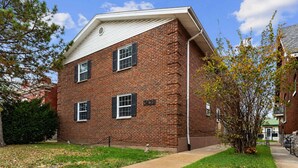 A two-story brick building with a flat roof, three windows with shutters, and a small front yard with grass and a tree.