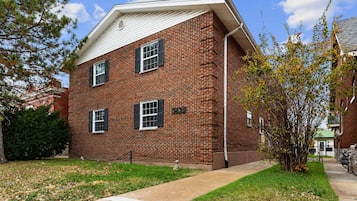 A two-story brick building with a flat roof, three windows with shutters, and a small front yard with grass and a tree.