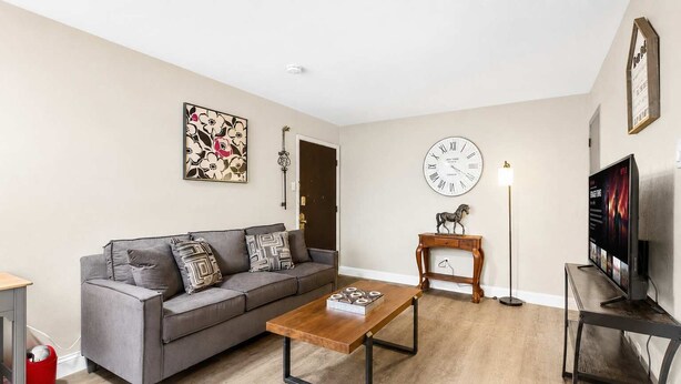 A living room with a grey sofa, a wooden coffee table, a clock, a TV, and a small wooden table with a horse figurine.