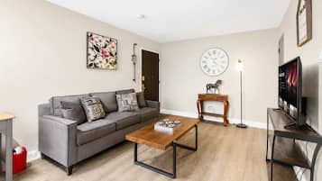 A living room with a grey sofa, a wooden coffee table, a clock, a TV, and a small wooden table with a horse figurine.