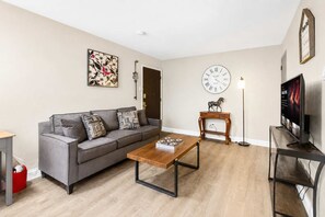 Ein Wohnzimmer mit grauem Sofa, einem Holztisch, einer Uhr, einem Fernseher und einem kleinen Holztisch mit einer Pferdefigur.
