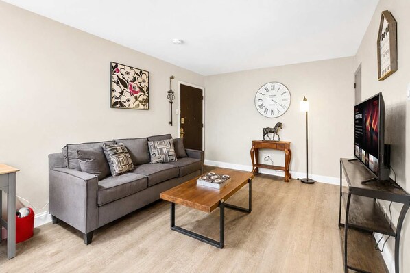 A living room with a grey sofa, a wooden coffee table, a clock, a TV, and a small wooden table with a horse figurine.