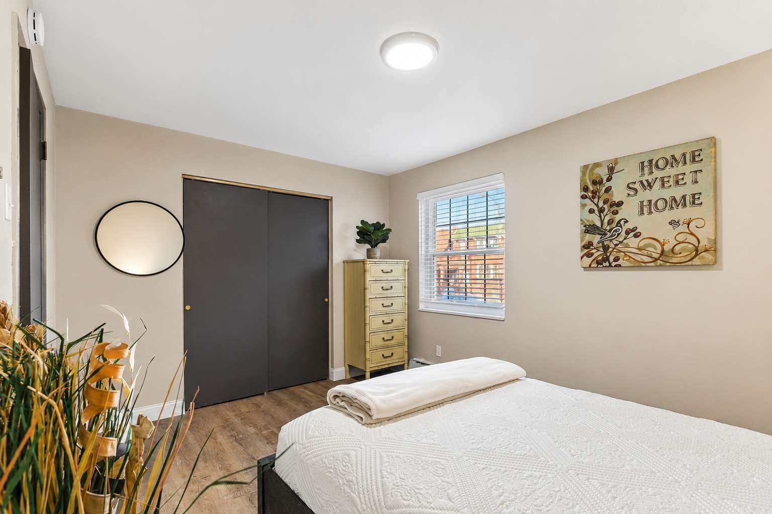 A bedroom with a bed, a dresser, a mirror, a window with blinds, and a wall decoration.
