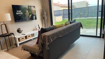 A modern living room with a sofa, a flat-screen TV, a lamp, and a view of a backyard.