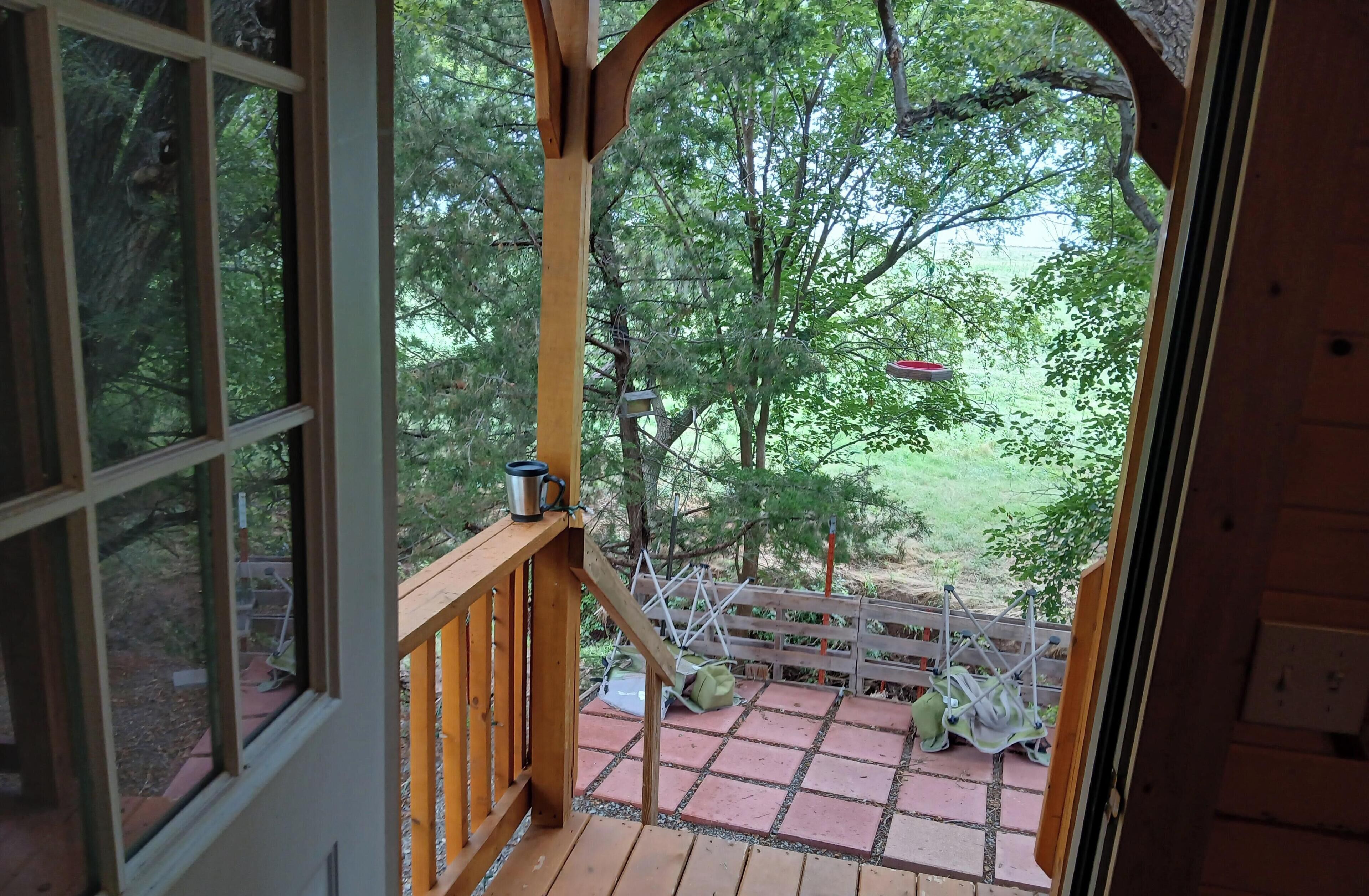 Une terrasse en bois offrant une vue sur une zone boisée, une mangeoire à oiseaux en métal et un sac vert.