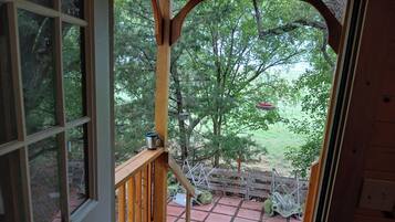 A wooden deck with a view of a forested area, a metal bird feeder, and a green bag.