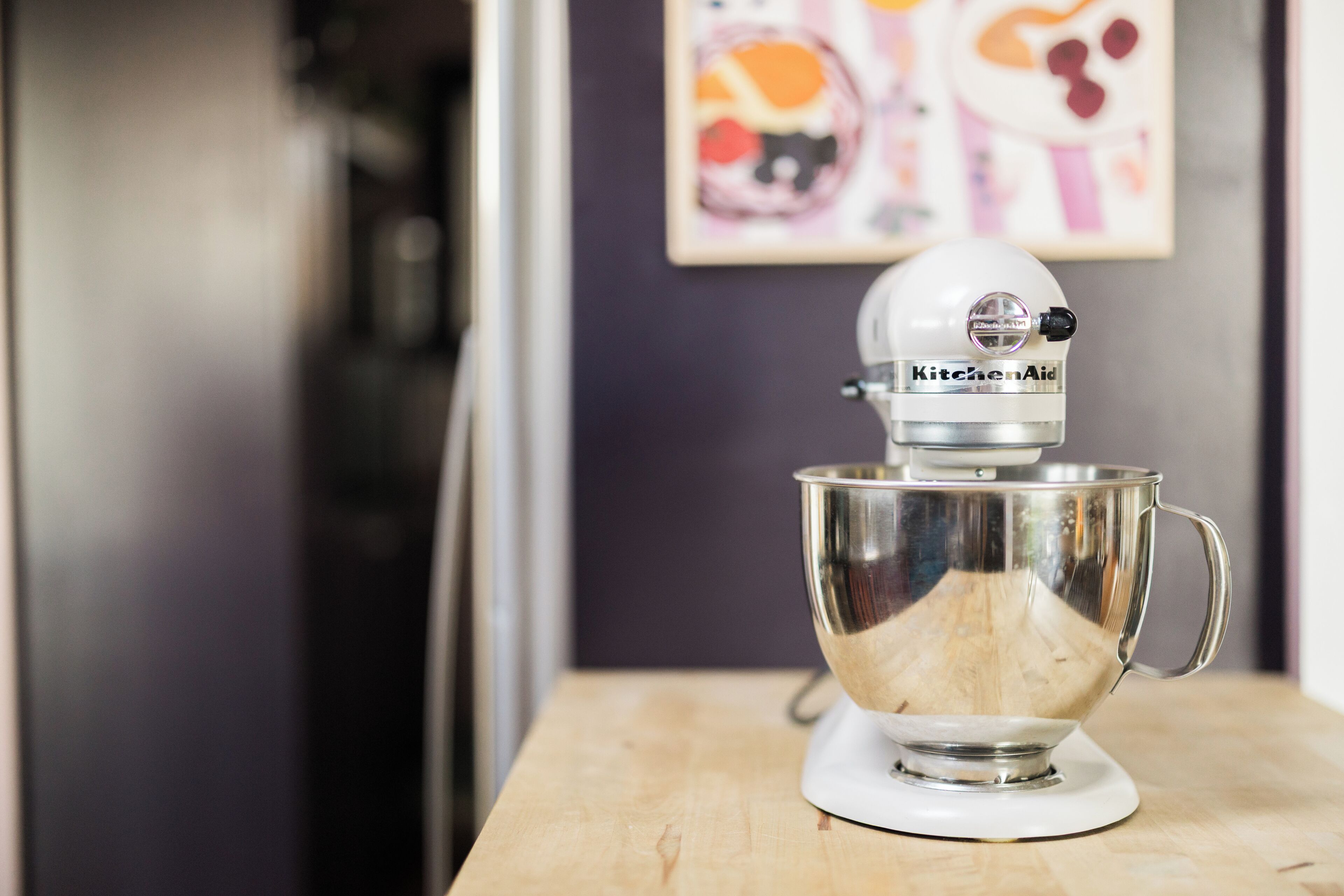 A KitchenAid stand mixer on a wooden table with a kitchen appliance in the background.