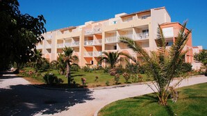 Un hotel de varios pisos con balcones, rodeado de palmeras y un jardín cuidado.