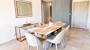 Un espace repas comprenant une table rectangulaire, des chaises blanches et un vase contenant des fleurs séchées.