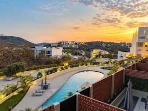 A swimming pool with a curved edge, surrounded by a patio area with chairs and umbrellas, and a view of a hillside with buildings in the background. - Casa Racél · Nirú Las Casitas · 2BR/2BA (Bahia de Santa Cruz)