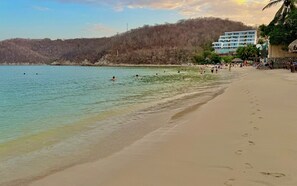 Ein Strand mit klarem Wasser, sandigem Ufer und einem mehrstöckigen Gebäude im Hintergrund.