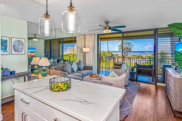 A modern living room with a marble island, a sofa, and a dining area with a view of the ocean. - Remodeled One BD Ocean, AC,wifi, pool, Luana Kai D202 (Kihei)