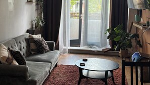 Een woonkamer met een grijze bank, een ronde salontafel, een bruin vloerkleed en een groot raam met gordijnen.
