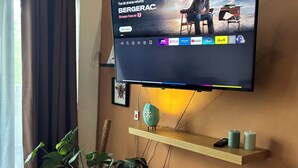 Een woonkamer met een tv aan de muur waarop een film wordt afgespeeld, een plant, een plank met kaarsen en een rieten mand.