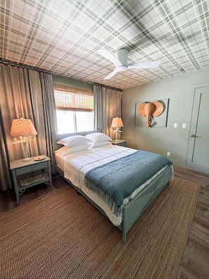 A bedroom with a bed, two bedside tables, a ceiling fan, a window with curtains, and a wall-mounted artwork.