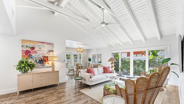 A spacious living room with a high ceiling, a white sofa, wicker chairs, a dining table, and a large window with a view.