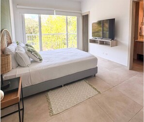 A bedroom with a bed, a TV mounted on the wall, a window with a view of trees, and a door leading to another room.