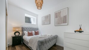 A bedroom with a grey tufted bed, a bedside table with a lamp, a window, and a white dresser. - Bon Accord Suite - Grampian Lettings (Aberdeen)