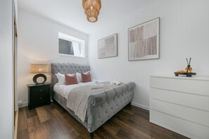 Ein Schlafzimmer mit einem grau gesteppten Bett, einem Nachttisch mit Lampe, einem Fenster und einer weißen Kommode.