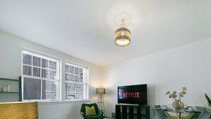 A living room with a television displaying the Netflix logo, a floor lamp, a green armchair, a dining table with chairs, and a large window with blinds. - Bon Accord Suite - Grampian Lettings (Aberdeen)