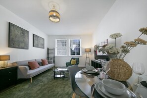 Una sala de estar moderna con un sofá, una mesa de comedor para dos personas, una mesita de centro y un jarrón decorativo con flores.