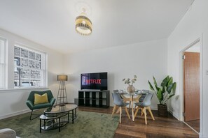 Una sala de estar con televisor, mesa de comedor, sillas, sofá, lámpara y planta en maceta.