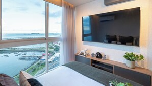 A modern hotel room with a large window, a bed, a TV, and a small plant. - Modern 1BR Ocean View | The Sands Pool High Floor (Panamá)