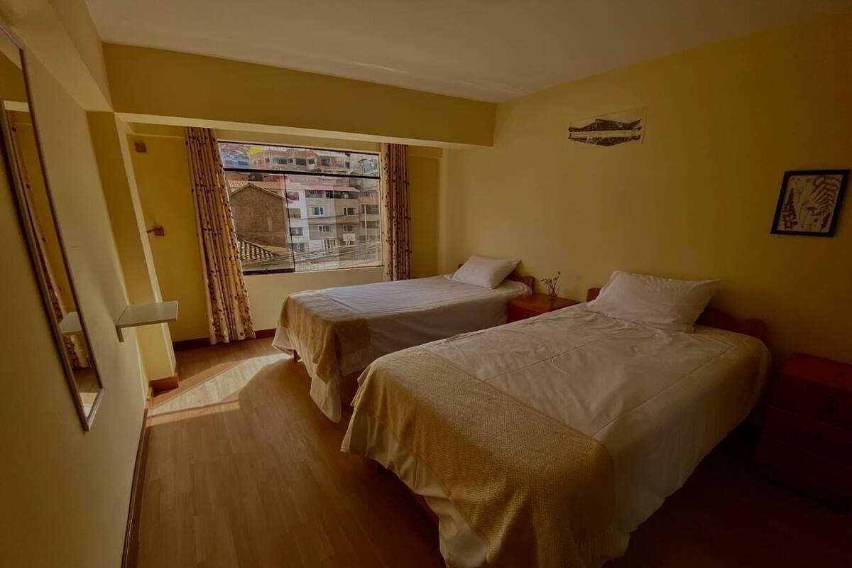 Ein Zimmer mit zwei Betten, einem mit Vorhängen versehenen Fenster und Blick auf Gebäude.