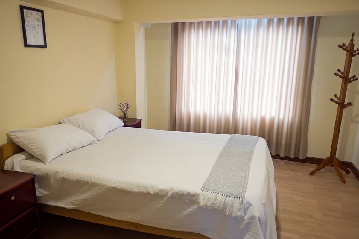 Ein ordentlich bezogenes Bett mit Kissen, ein Nachttisch mit einer Vase und ein Kleiderständer in einem Zimmer mit Fenster und Vorhängen.