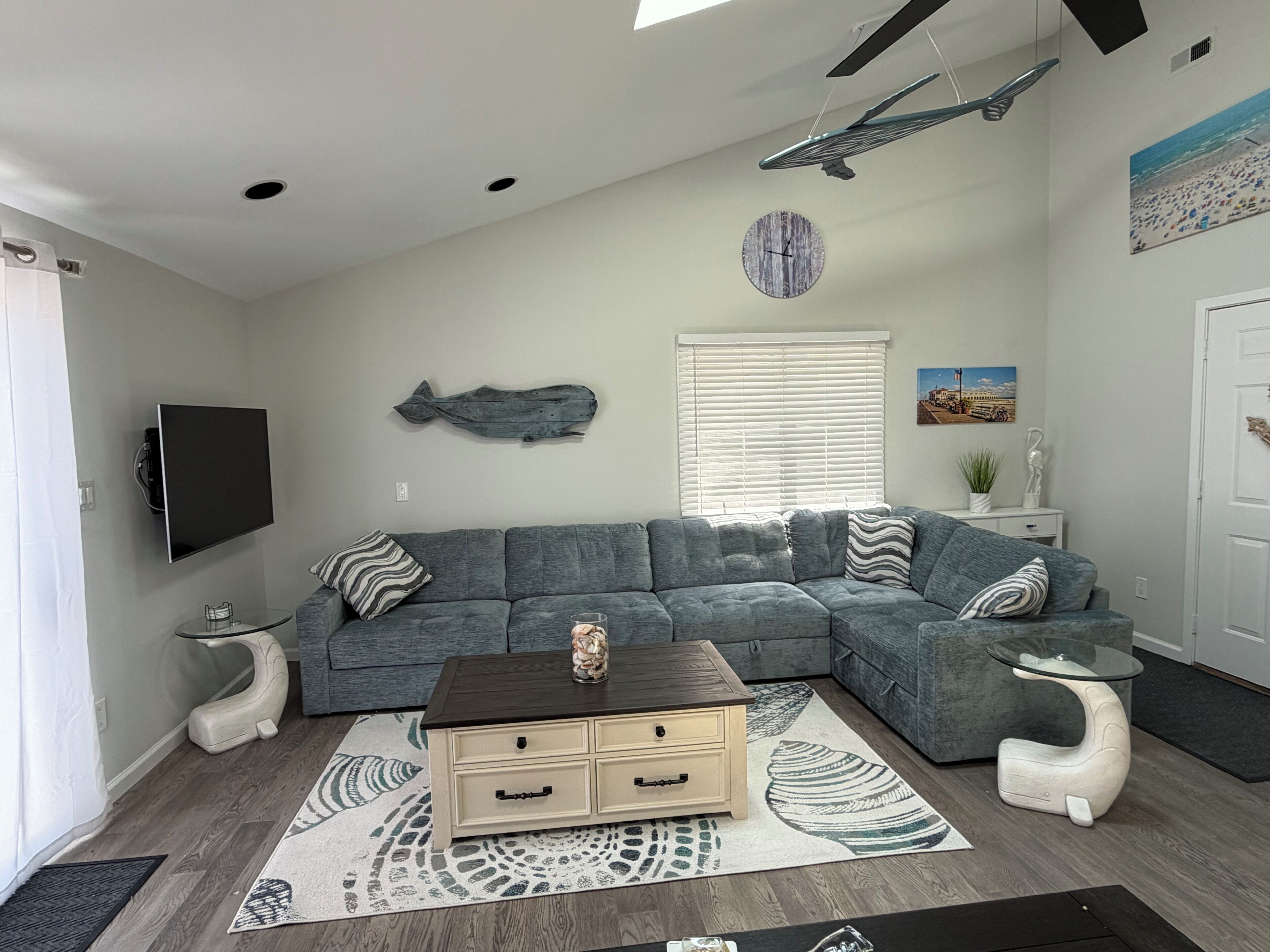 Una sala de estar con un sofá modular, una mesa de centro, un televisor de pantalla plana, un reloj de pared y un ventanal con persianas.