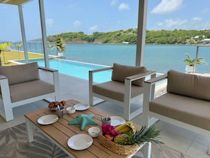 A poolside area with two sofas, a table with plates, cups, and a fruit basket, overlooking a clear blue sea and lush greenery. - Villa Caribbean Tradewinds, exceptional sea view (Le Vauclin)