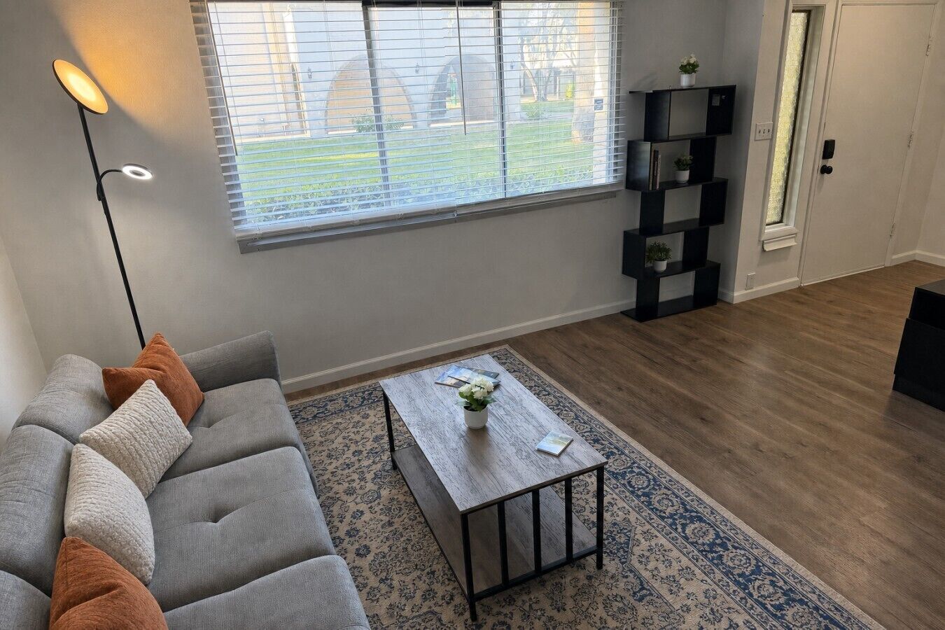 A living room with a grey sofa, a coffee table, a floor lamp, a window with blinds, and a bookshelf.