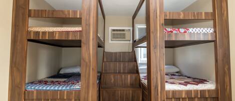 Een kamer met drie houten stapelbedden, een trap in het midden en een klein raam met airconditioning.
