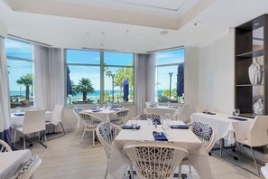 Ein Restaurant mit weißen Tischen und Stühlen, große Fenster mit Blick auf das Meer und Palmen.