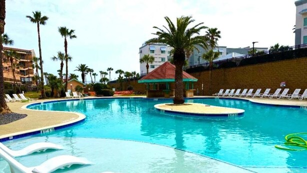 A swimming pool with lounge chairs, palm trees, and a small building with a red roof. - Ocean View, Sea Ya Next Tide – 2 BR/2 BA - Sleeps 6 (Galveston)