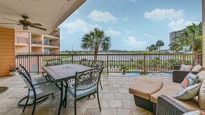 A balcony with a table and chairs, a sofa, a ceiling fan, and a view of a body of water and palm trees. - Ocean View, Sea Ya Next Tide – 2 BR/2 BA - Sleeps 6 (Galveston)