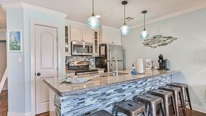 A kitchen with a central island, bar stools, a microwave, oven, and a sink. - Ocean View, Sea Ya Next Tide – 2 BR/2 BA - Sleeps 6 (Galveston)