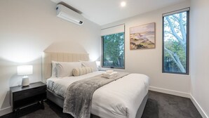 Une chambre à coucher comprenant un lit, une table de chevet, une lampe, un appareil de climatisation, de grandes fenêtres et des œuvres d’art.