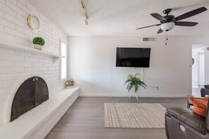 Ein Wohnzimmer mit Kamin, einem flachbildfernseher an der Wand, einem Deckenventilator und einem Blumentopf.