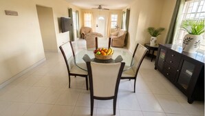 Uma sala de jantar com mesa de vidro rodeada por cadeiras, uma cesta de frutas e um armário escuro com objetos decorativos.