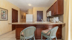 A kitchen with wooden cabinets, a refrigerator, a microwave, a coffee maker, and two wicker chairs. - Sonoran Sun SW-414 Ocean Front Wraparound Terrace Condo (Puerto Peñasco)