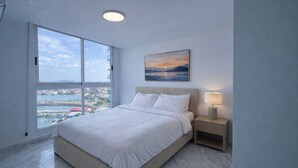 Et soveværelse med seng, et natbord, en lampe, et maleri og et stort vindue med udsigt over byen.