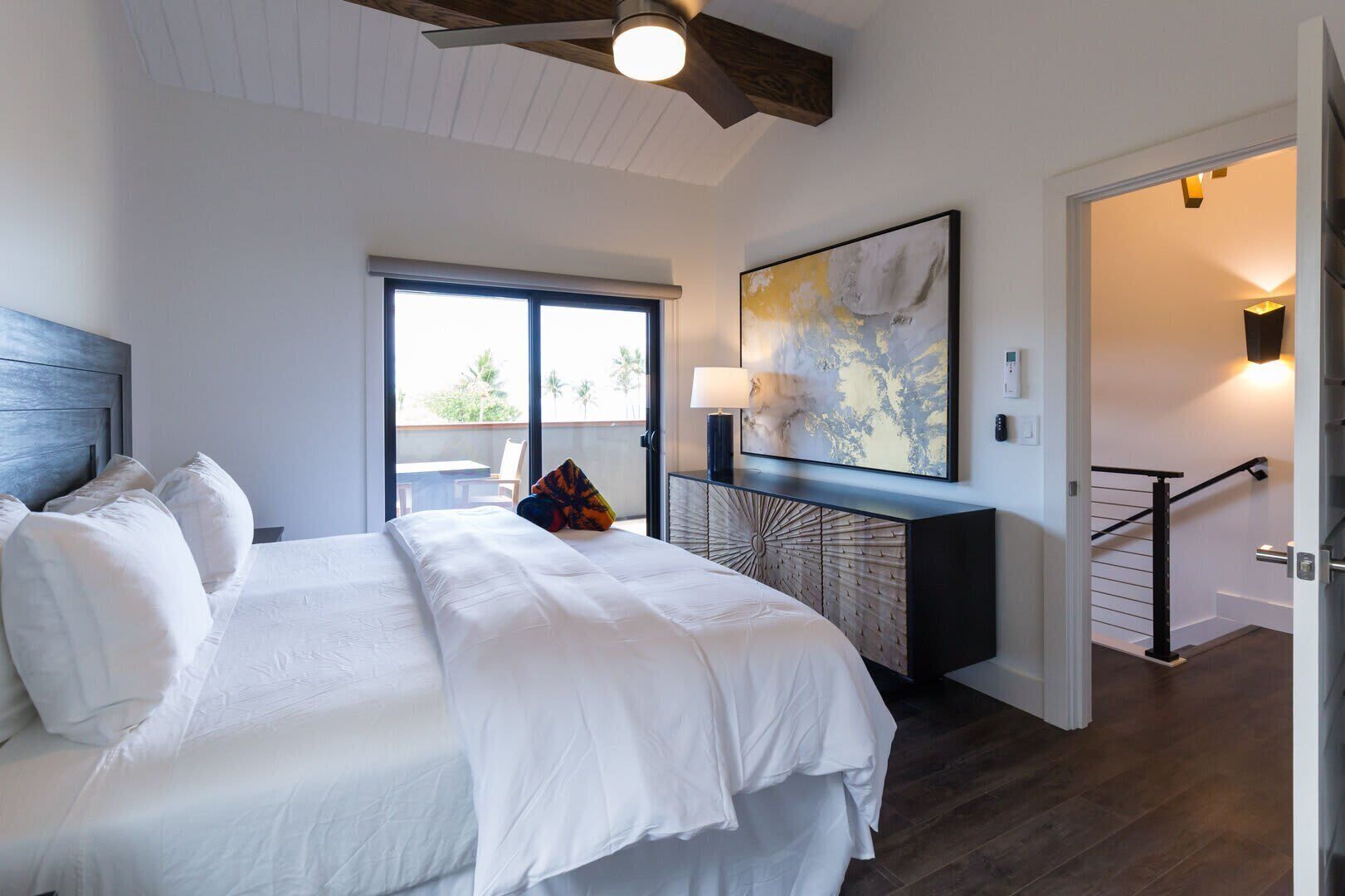 A bedroom with a large bed, a desk with a lamp, a painting, and a view through a sliding door.