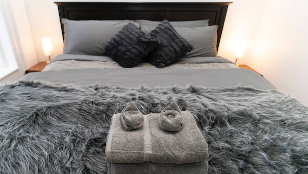 A bed with a dark headboard, grey bedding, and a fluffy grey rug.