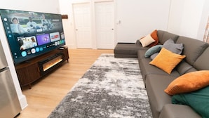A living room with a flat-screen TV, a grey sofa with colorful cushions, a patterned rug, and a wooden entertainment unit.