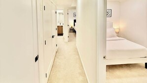 A hotel corridor with white walls, a carpeted floor, and a bed with a headboard in the right room.