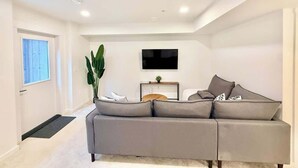 A modern living room with a grey sofa, a flat-screen TV mounted on the wall, a small wooden side table, and a large green plant.