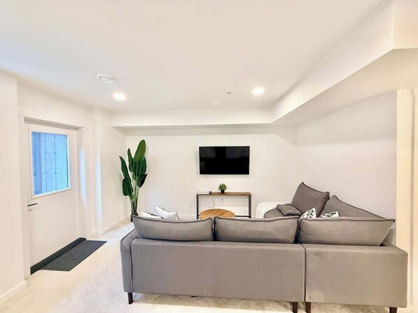 A modern living room with a grey sofa, a flat-screen TV mounted on the wall, a small wooden side table, and a large green plant.